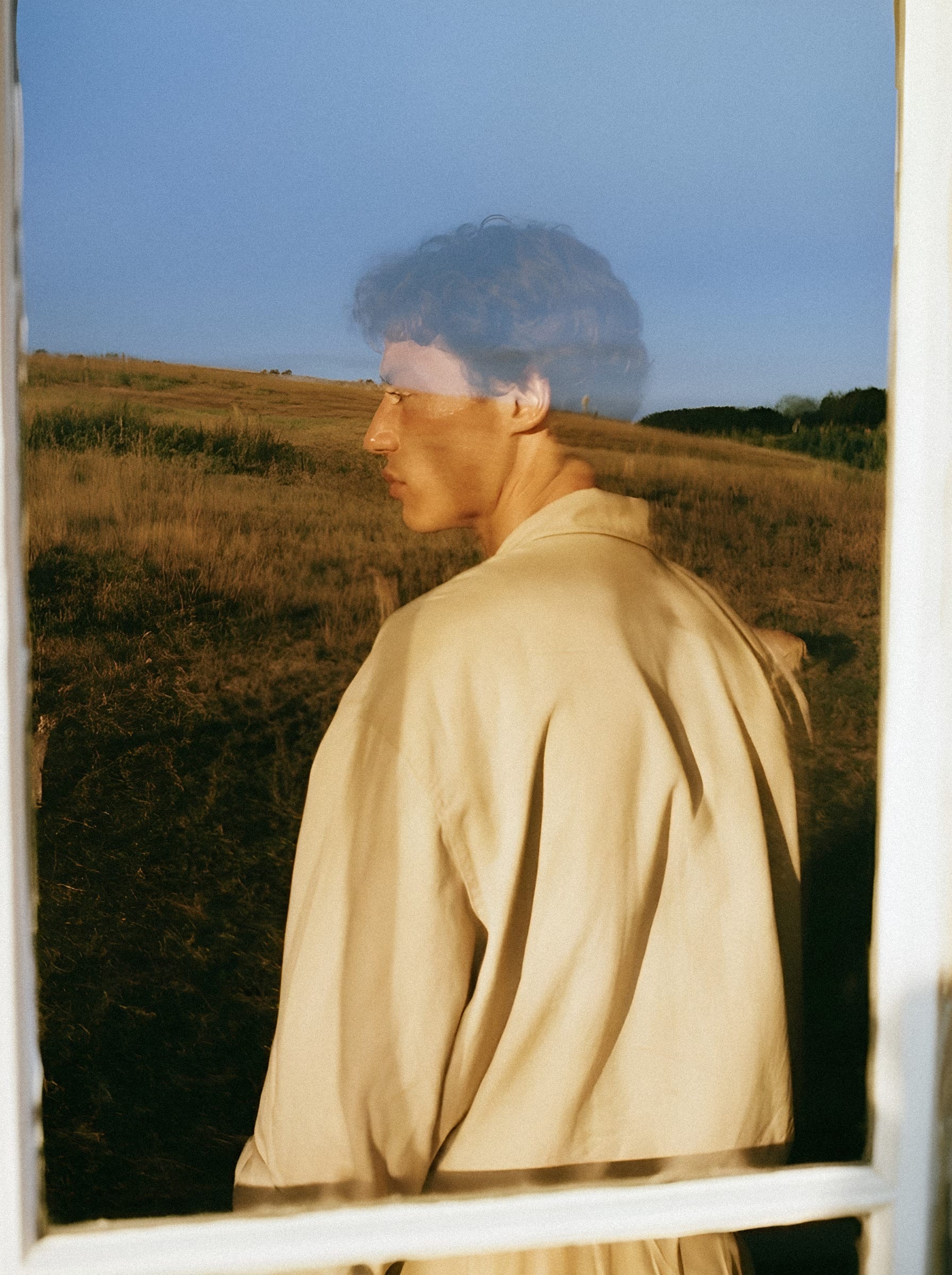 A man in a linen shirt reflected in a window overlooking grassland.