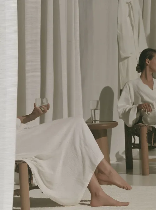 Two seated figures in white linen beside a sunlit curtain and glasses of water.